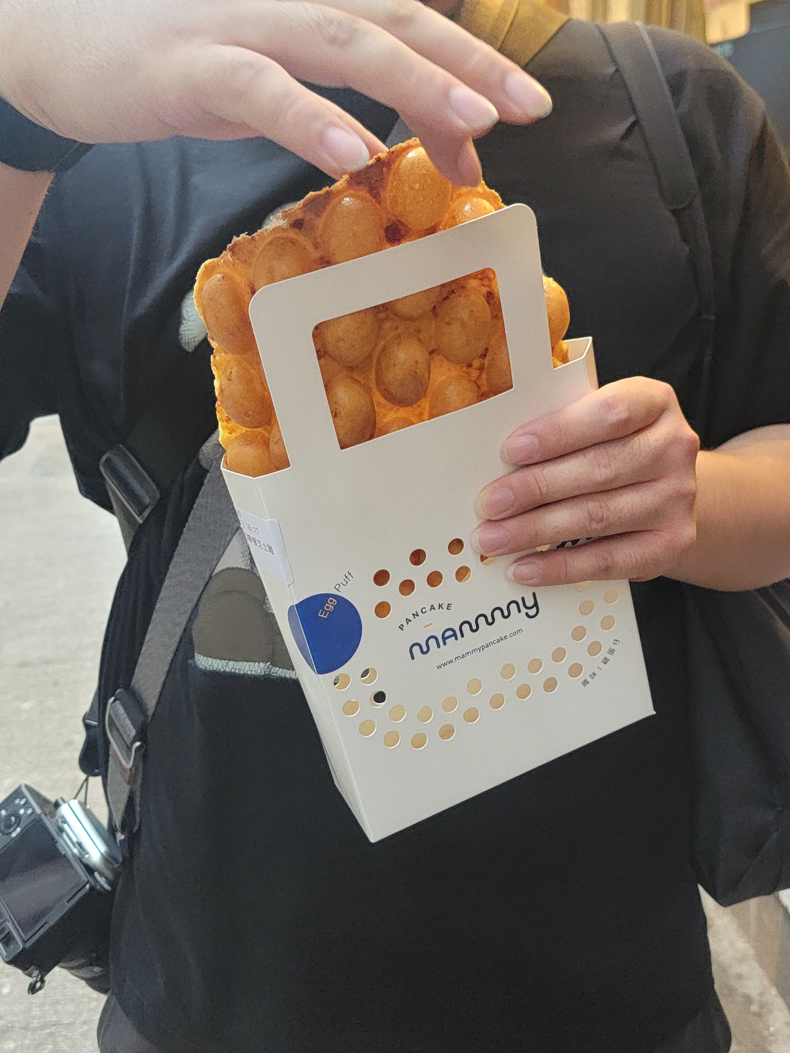 Hand holding a Hong Kong egg waffle in a paper sleeve.